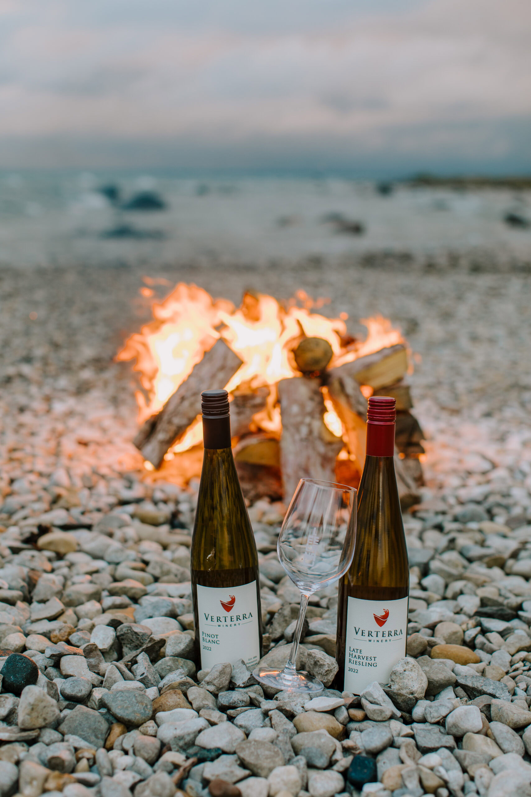 Two bottles of Verterra wine and a glass on a rocky beach with a bonfire in the background.